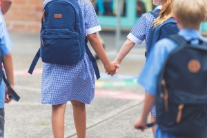 Varios niños dados la mano se encaminan al colegio con u mochila y un babi
